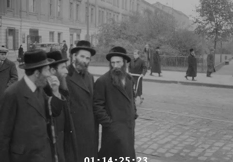 Four men in long coats walking beside trees, with more people on the far sidewalk, Kraków, 1936