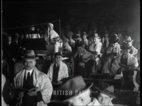 British Pathé frame: crowded hall of men in hats and tallisos, bimah in the background