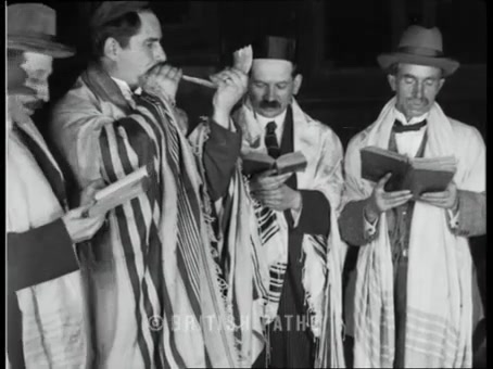 British Pathé frame: man in tallis blowing a shofar beside three other men holding open prayer books