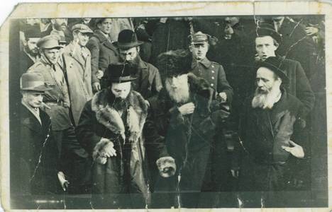 Known photograph of Reb Pinyele Ostiler walking with two men holding his arms, in a fur hat and dark coat