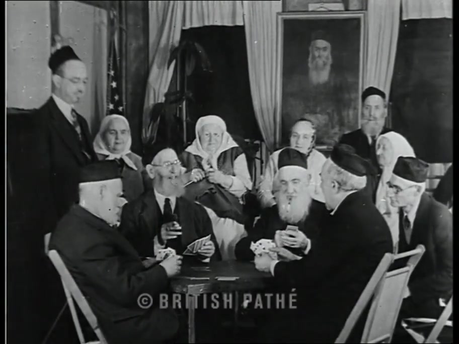 British Pathé frame: elderly men in yarmulkes playing cards at a table, women watching, American flag and portrait on the wall