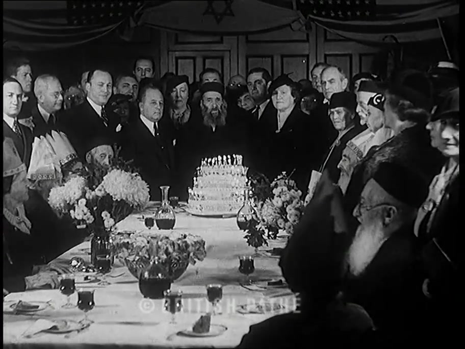 British Pathé frame: birthday cake with many candles, Magen David banner and American flags, elderly bearded man at center