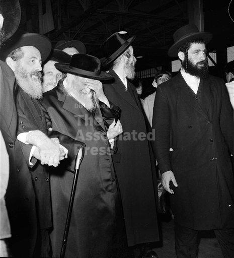 The Satmar Rebbe, Reb Yoel Teitelbaum, at the Queen Mary pier on August 31, 1965, hand over his face, with men in black hats under the pier roof