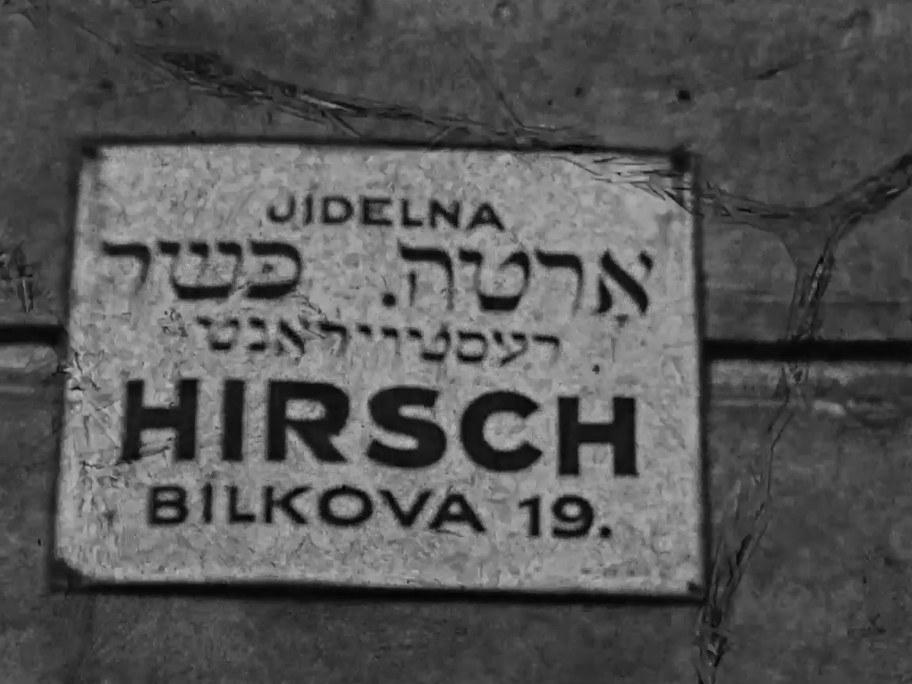 1934 film frame: Jídelna Hirsch kosher restaurant sign, Bílkova 19, in Czech, Hebrew, and Yiddish