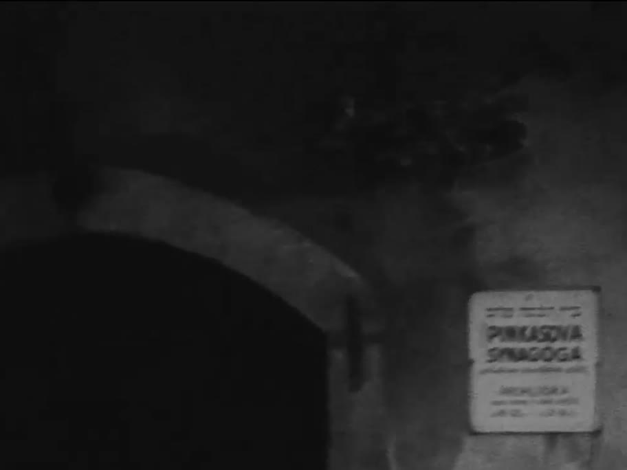 1936 film frame: wall sign reading PINKASOVA SYNAGOGA and PROHLÍDKA beside a stone arch
