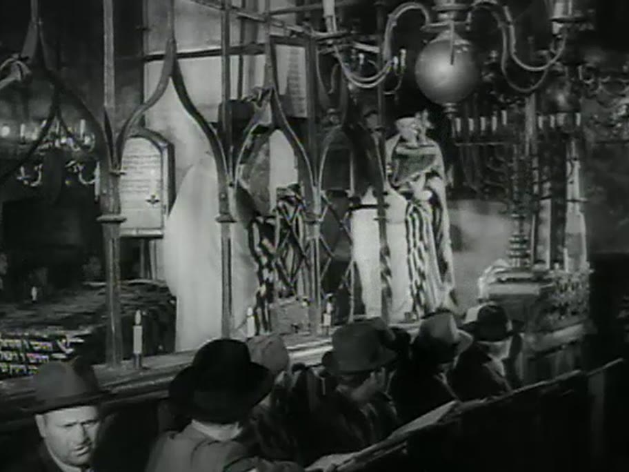 1946 film frame: Torah ceremony inside the Old-New Synagogue, wrought-iron bimah cage, men in hats and tallisim